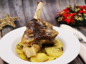 Un codillo de cordero asado sobre una base de patatas a lo pobre en un plato blanco sobre una mesa de madera con decoración de Navidad